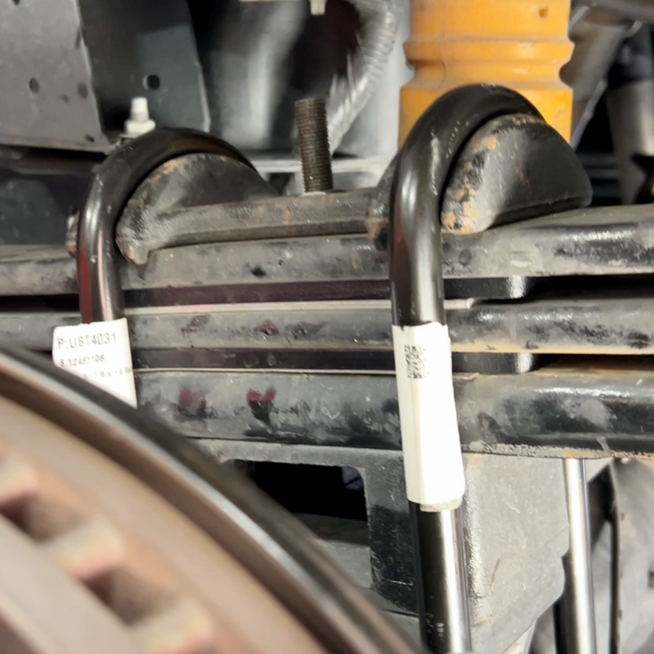 A set of metal leaf spring shims placed under a vehicle's leaf spring, with center and roll pins visible.