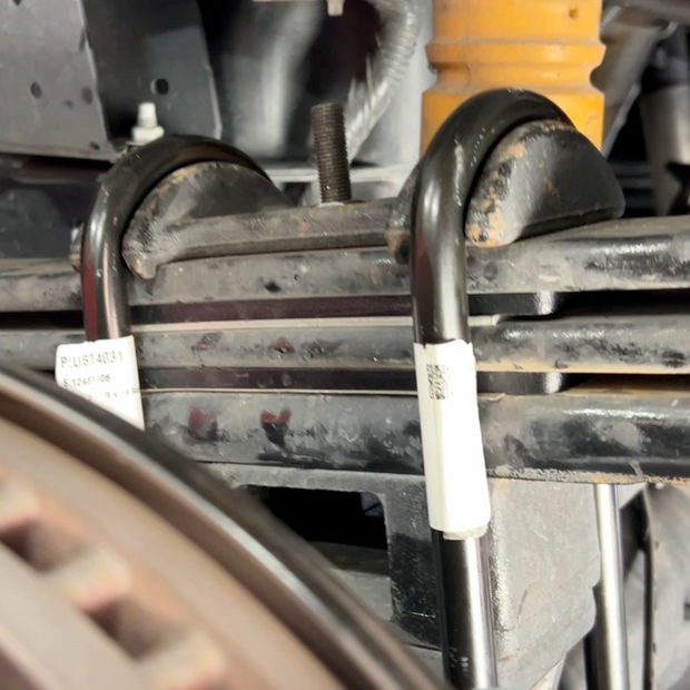 A set of metal leaf spring shims placed under a vehicle's leaf spring, with center and roll pins visible.