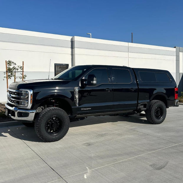 A black 2023+ Ford F250/F350 truck with a 2.5 inch lift kit, showcasing the raised front and the new aftermarket wheels and tires.